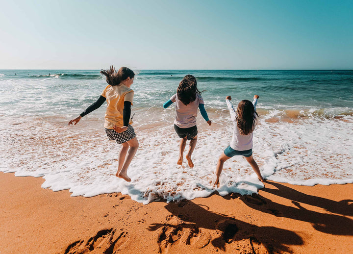 Three girls excitedly jumping over the waves on the shoreline, wearing Crazy Arms sun sleeves
