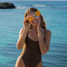 Woman in a brown swimsuit holding a sunbutter sunscreen in front of her face with a blue ocean background