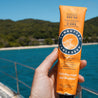 Hand holding a Lushutu Fun Sunscreen tube with a scenic background of water and trees.