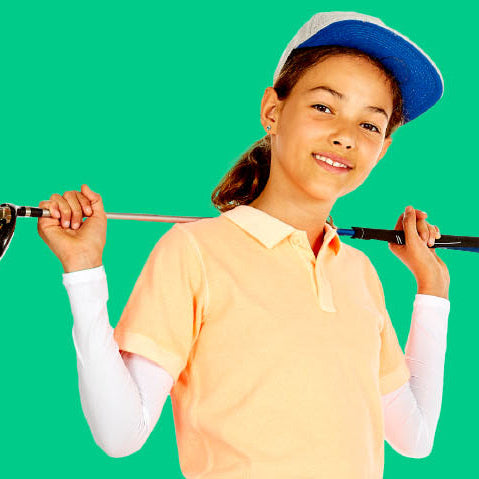 A young girl smiling and holding a golf club over her shoulder, wearing a peach polo shirt, white long-sleeve undershirt, blue and white cap, and white arm sleeves, set against a solid bright green background.